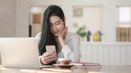ノートパソコンとコーヒーを前に、スマートフォンを見ながら笑顔を見せる外国人女性の写真